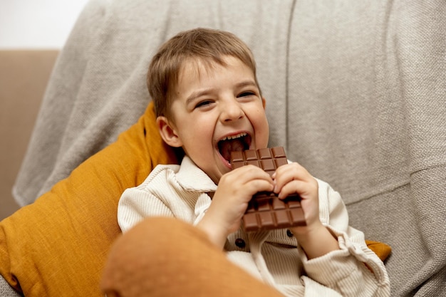 Little adorable boy sitting on the couch at home and eating chocolate bar Child and sweets sugar confectionery Kid enjoy a delicious dessert Preschool child with casual clothing Positive emotion