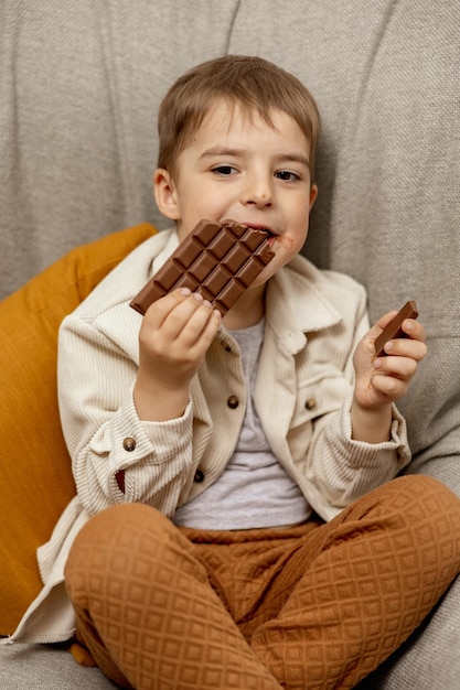 家のソファに座ってチョコレートバーを食べている小さな愛らしい男の子子供とお菓子の砂糖菓子子供はカジュアルな服装でおいしいデザート就学前の子供を楽しんでいますポジティブな感情