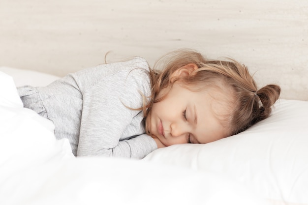 Little adorable child girl in grey pajamas closed eyes lying in bed sleeping on comfortable pillow and under white fluffy cotton blanket, good night sweet dreams