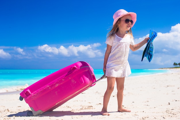 Little adorable girl with big luggage in hands on tropical beach