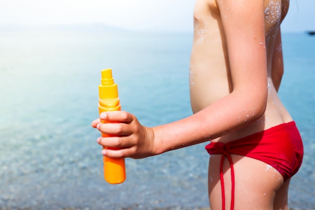 Piccola ragazza adorabile con la bottiglia di crema solare sulla spiaggia