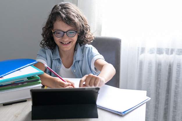 Piccolo adorabile ragazzo di scuola con gli occhiali che studia online da casa bambino utilizzando la tavoletta digitale