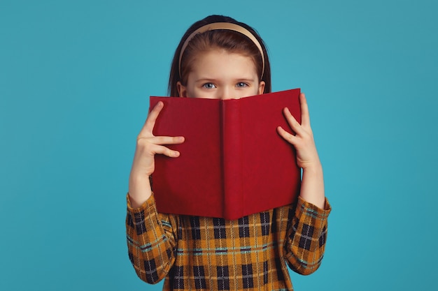 Photo little adorable schoolgirl hides behind book has joyful expression