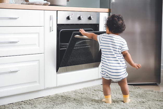 Little African-American baby near stove in kitchen. Child in danger