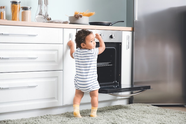 Little African-American baby near stove in kitchen. Child in danger