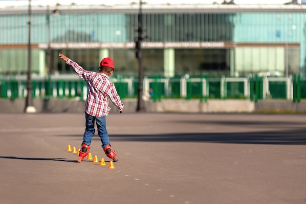 롤러 스케이트를 탄 어린 아프리카계 미국인 소년은 훈련 원뿔 사이에서 아스팔트 도로를 스케이트를 탄다.