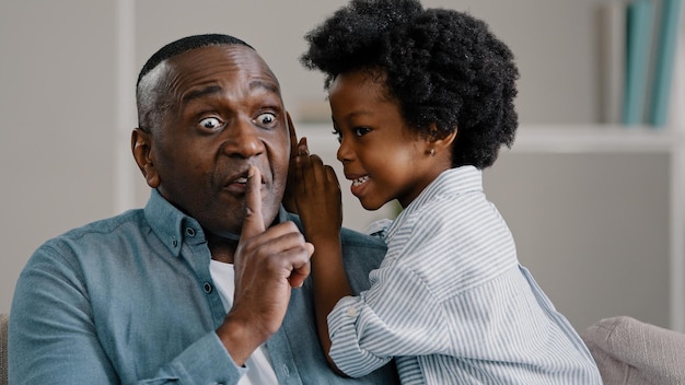 Little african american daughter whispering secret adult dad in ear telling gossip surprised dad