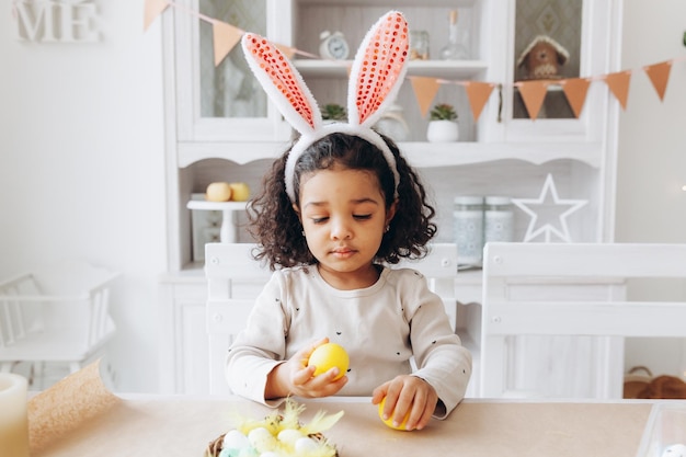 Little African American girl paints Easter eggs at home in the kitchen happy easter