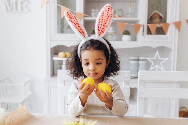 Little African American girl paints Easter eggs at home in the kitchen happy easter