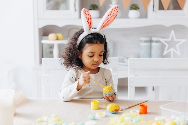 Little African American girl paints Easter eggs at home in the kitchen happy easter