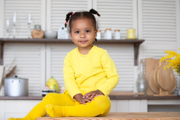 A little African American girl in a yellow dress with curly pigtails sits in the kitchen