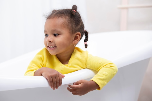 Little African American girl in a yellow dress with curly pigtails sits in a retro bath.