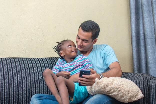 a little african girl or daughter sitting on a sofa and on the laps her white father