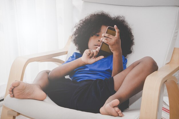 Little African kid is playing with smartphone on sofa