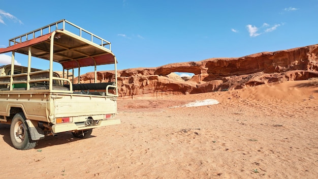 Little arc or small rock window formation in Wadi Rum desert, blue sky above, rear of vehicle for driving tourists in foreground