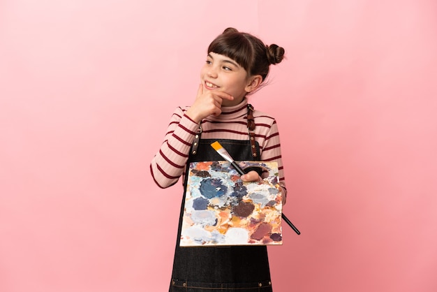 Little artist girl holding a palette isolated on pink looking to the side and smiling