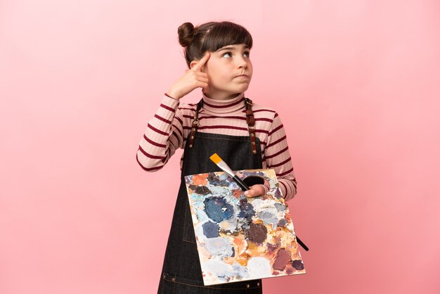 Little artist girl holding a palette isolated on pink wall having doubts and thinking