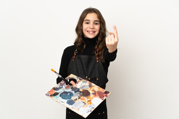 Little artist girl holding a palette isolated on white background doing coming gesture