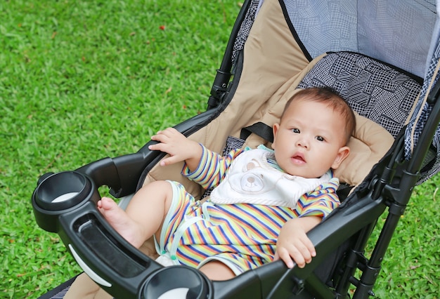 Маленький азиатский мальчик сидит в коляске в зеленом саду.
