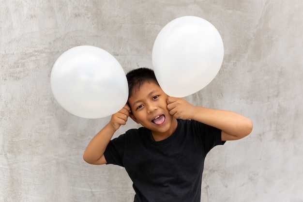 Little asian boy holding white balloons over grey concrete background.