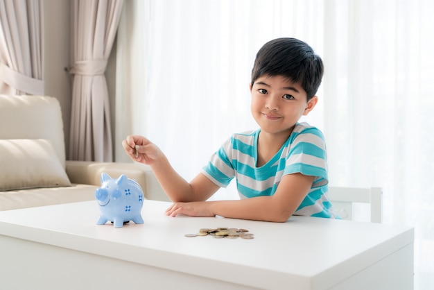 Little Asian boy insert a coin into blue piggy bank in white table at living room at home for the kid saving.