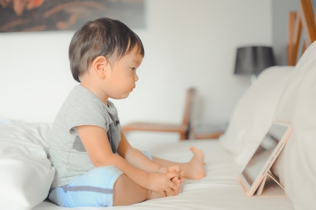 Little asian boy lying in bed and watches a story on touchscreen tablet