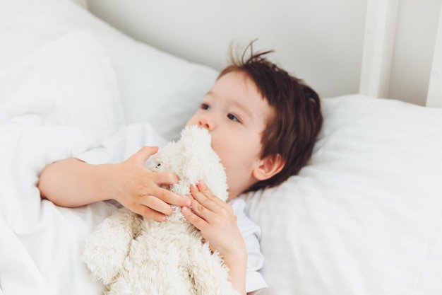 A little Asian boy in pajamas lying on the bed with a teddy bear The child wakes up happy The child wakes up early in the morningnatural bed linen for children