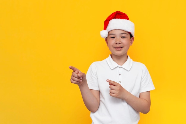 little asian boy in santa hat shows his hands to the side on yellow isolated background korean child