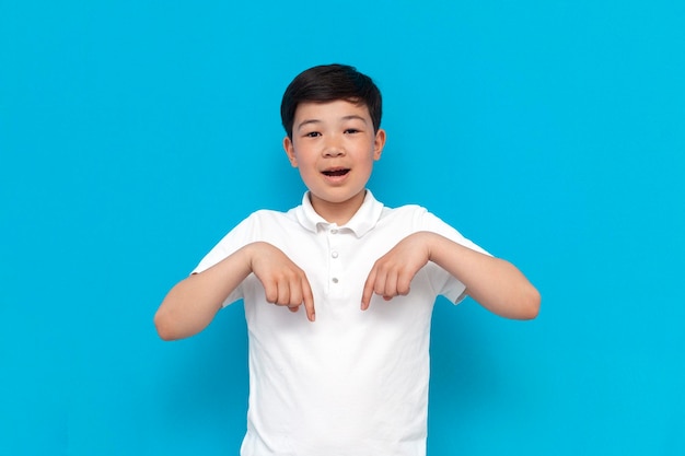 Little asian boy in white tshirt pointing down on blue background korean kid advertising copy space