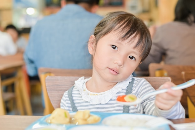 レストランのテーブルで昼食を食べている間不幸な顔を持つ小さなアジアの子女の子、つまらない食べる人は食べたくないまたは空腹ではない