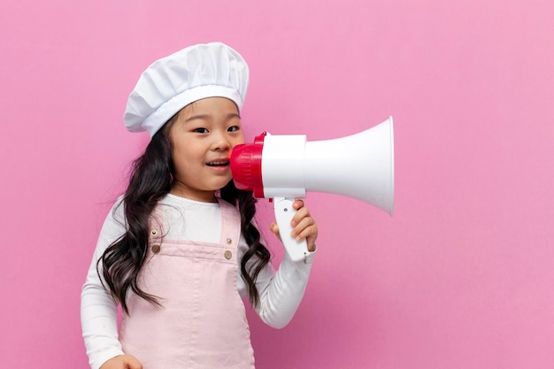 little Asian girl in chef uniform announces news and information with megaphone on pink background