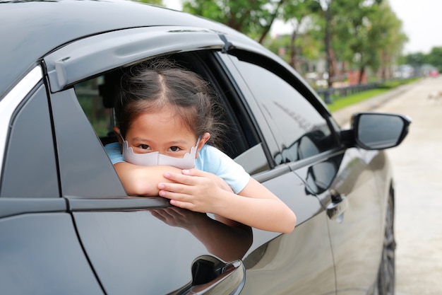 Little Asian girl child wears hygiene face mask looking through camera with pokes her head out of car window