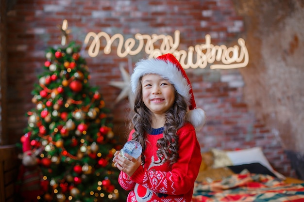 Photo little asian girl holding a glass snow globe on the background of a christmas tree christmas