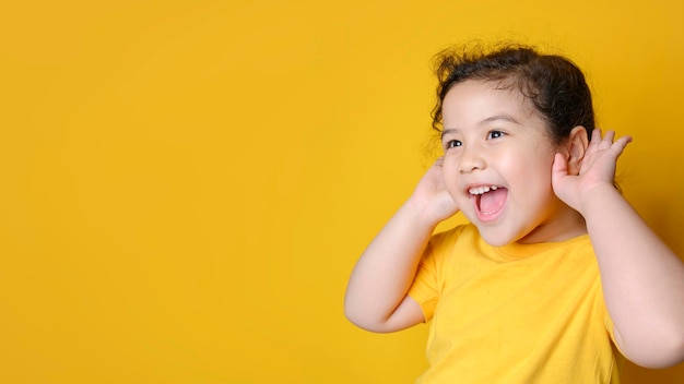 La piccola ragazza asiatica tiene la mano vicino all'orecchio e ascolta. faccia emozionante su una bambina asiatica. faccia eccitante su una ragazza asiatica felice che indossa una camicia gialla e ascolta curiose buone notizie.