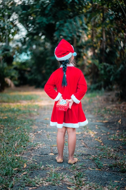 写真 赤いサンタクロースの衣装を着た小さなアジアの女の子が公園のプレゼントボックスで後ろに立ちます