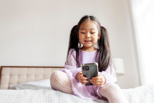 작은 아시아 소녀는 집에서 침대에 앉아 스마트폰을 사용하고 있습니다. 아이는 전화를 보고 있습니다.