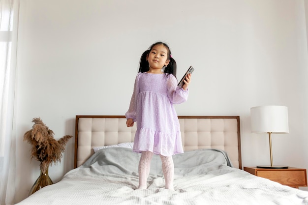 little asian girl jumping on the bed at home and using smartphone the child looks at the phone