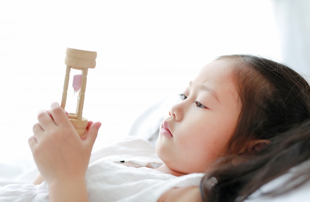 Little Asian girl looking at hourglass in hand lying on bed at home.