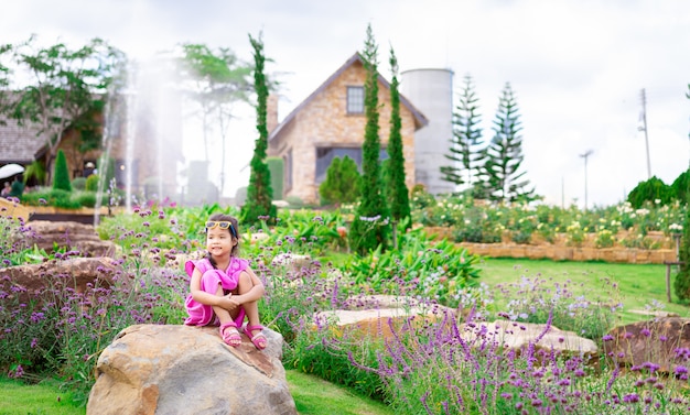 Маленькая азиатская девушка в розовом платье, сидя на скале в цветочном саду