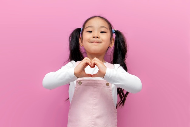 little Asian girl in pink sundress with long hair shows heart with her hands Korean preschool child