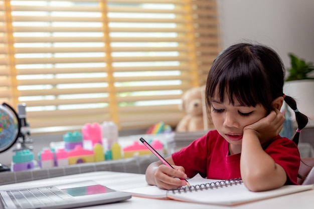 Little Asian girl sitting alone and looking out with a bored face Preschool child laying head down on the table with sad bored with homework spoiled child