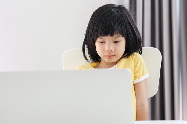 小さなアジアの女の子の学生は、自宅でラップトップコンピューターを使用してオンラインで勉強します