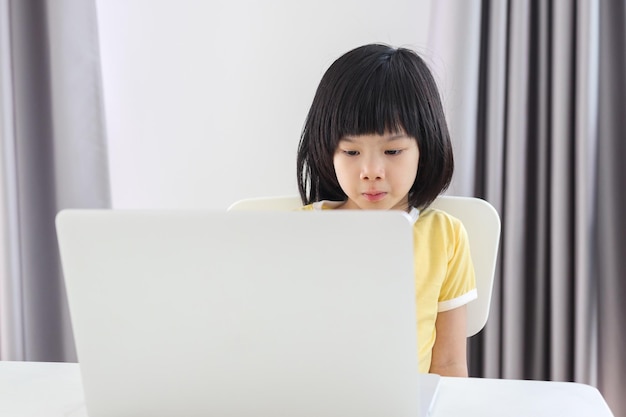 小さなアジアの女の子の学生は、自宅でラップトップコンピューターを使用してオンラインで勉強します