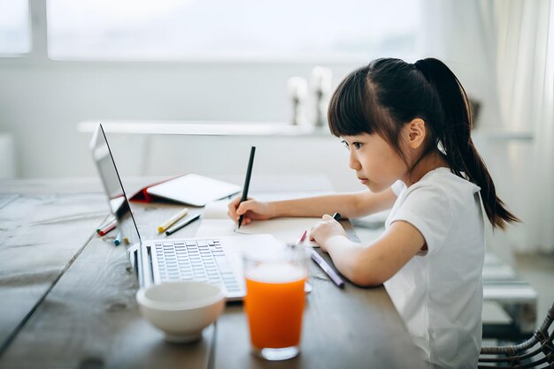 Little Asian girl studying from home She is attending online school classes with laptop and digital tablet and writing notes at home