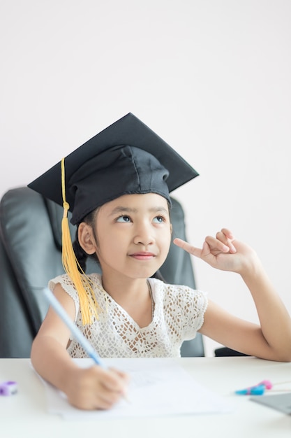 Little Asian girl wearing graduate hat doing homework 