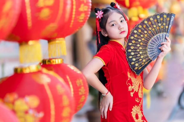 写真 赤い伝統的な中国のチャイナドレスを着て、それに中国のアルファベットの祝福が書かれたファニングとランタンを持っている小さなアジアの女の子は、新年の幸運の祝福の褒め言葉の装飾です