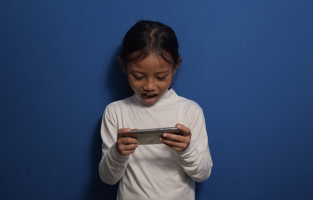 Маленькая азиатская девушка в белой футболке улыбается и смотрит на смартфон с удивленным и серьезно лицом