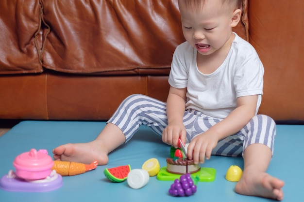 Маленький азиатский малыш сидит дома на полу и весело играет в одиночестве с кулинарными игрушками
