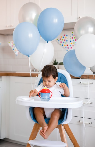 Piccolo neonato seduto nel seggiolone blu a casa sulla cucina bianca e acqua potabile dalla tazza sippy su sfondo con palloncini.