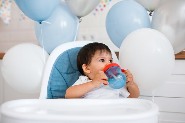 Little baby boy sitting in blue high chair at home on white kitchen and playing with wooden big spoon